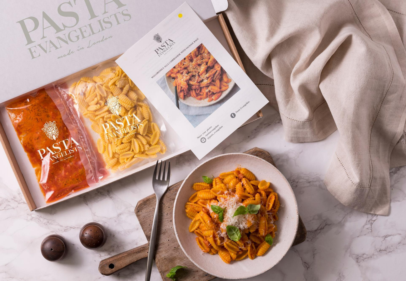 An overhead view of a Pasta Evangelists subscription box containing vacuum-sealed fresh pasta and sauce, alongside a recipe card and a finished bowl of pasta garnished with basil.